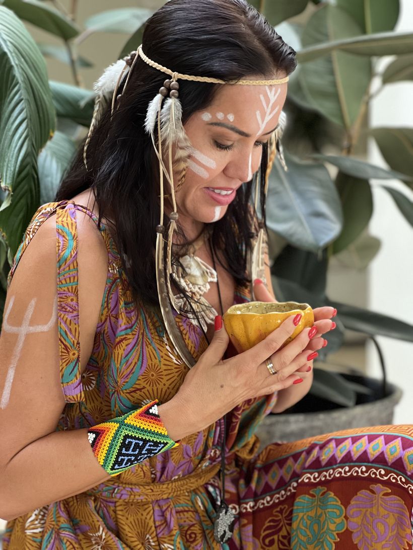 Márcia holding a cup of cacao, with white face paint and feathers in her hair