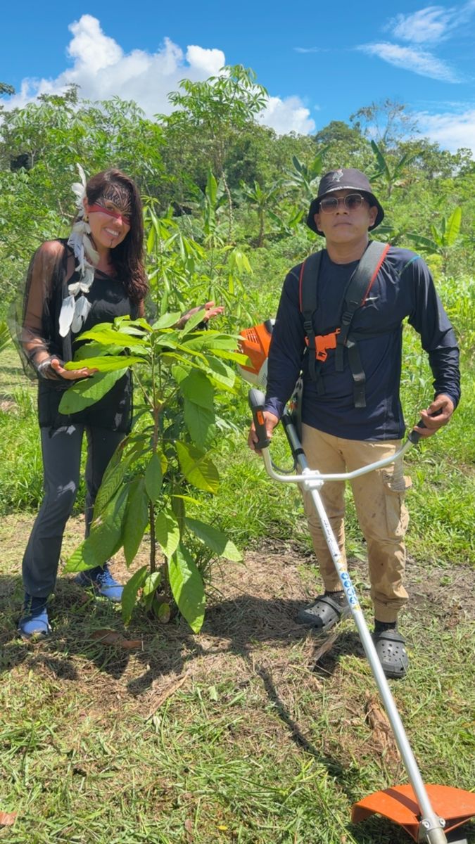 Márcia beside a young cacao tree, next to a community member holding a brush cutter