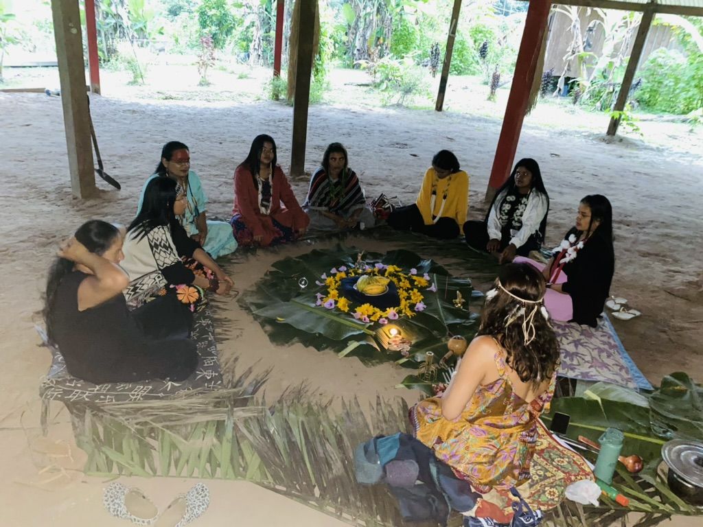 Yawanawá women sitting in a circle around an altar of leaves and yellow flowers, with Márcia among them