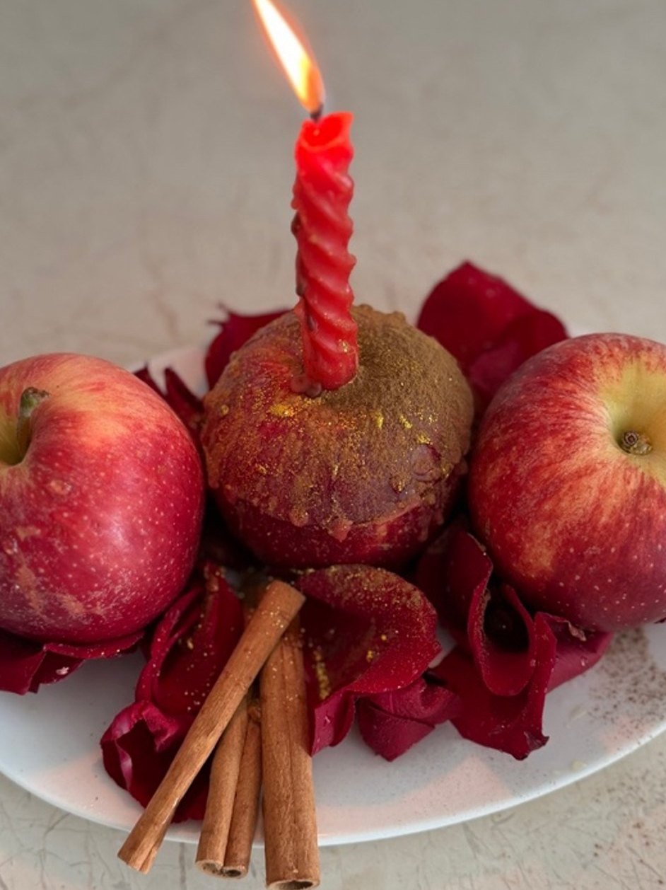 An apple sealed with red wax and covered in honey, a red candle burning on top, with rose petals, cinnamon sticks and two apples beside it
