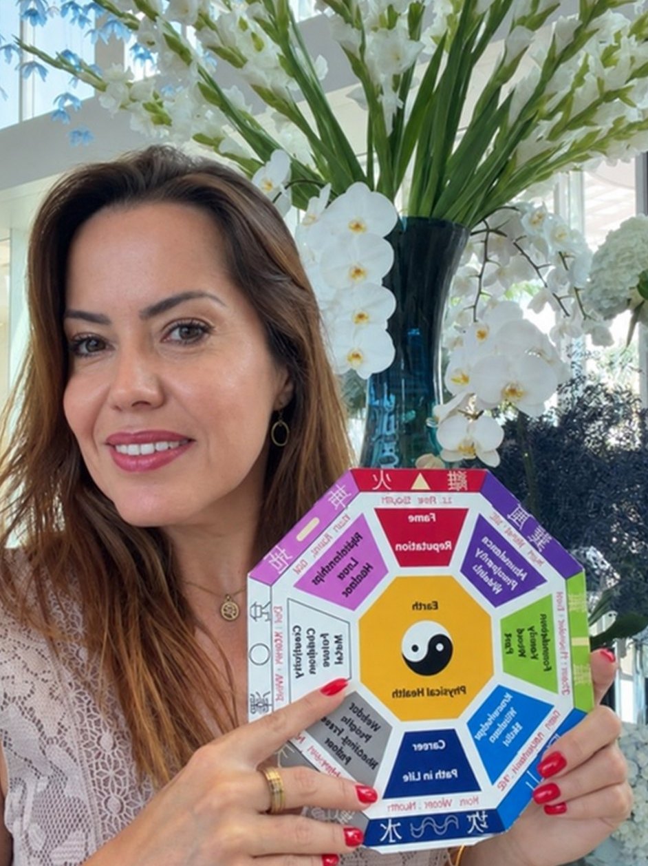 Márcia holding a colourful Baguá chart in front of a vase of white flowers