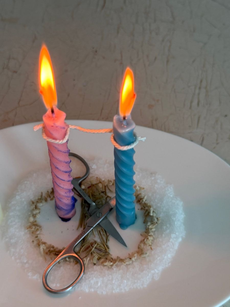 Two twisted candles tied together with a cord on a plate of salt and herbs, scissors between them
